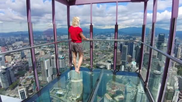 一个女人赤脚站在玻璃桥上无所畏惧 玻璃悬挂在吉隆坡天际线上方 俯瞰着这座城市高耸的大楼和繁华的街道 令人叹为观止 — 图库视频影像