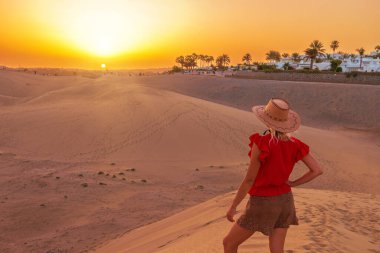 Gün batımında Gran Canaria 'nın Maspalomas Kumulları' nda güneş şapkası takan bir kadın turist. Ya uzak okyanus dalgalarını dinlemek için gözlerini kapatır ya da rüzgarda savrulan kum tepelerini seyreder..