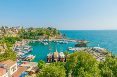 Türkiye 'nin Antalya kentinin panoramik manzarası. Akdeniz 'in derin mavi-yeşil suları çeşitli boyutlarda teknelerle dolu hareketli bir limana rastlar. Beyaz bir deniz feneri kayalık bir çıkıntıda nöbet tutuyor.