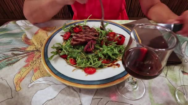 カラフルな花のテーブルクロスの背景に設定された 豊かな赤ワインのグラスとペアリングされたプレートに中希少なステーキサラダの活気に満ちた饗宴を食べる女性 サラダと赤ワインを提供 — ストック動画