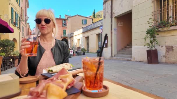 イタリアのエミリア ロマーナ地域の歴史的な村 ブリッジレラの中心部でカクテルを楽しむ観光ガール 生のハムと三日月のピエナ ロマニョーラをフィーチャーした食前酒にふけるシェイクスピア — ストック動画