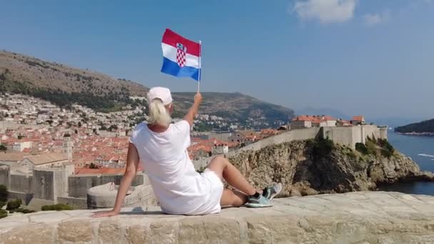 スローモーション クロアチアのドゥブロヴニクのラブリエンク砦の上にクロアチアの旗を持つ女性 ウエストハーバーと市壁の様子 ユネスコの都市ドゥブロヴニクは ダルマチアのクロアチアの古いベネチアの町です — ストック動画