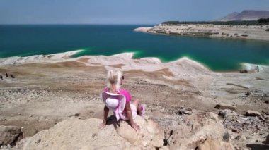 Düşünceli genç kız bir kayanın üzerinde oturup durgun ölü deniz sularına ve Ürdün 'deki açık mavi gökyüzünün altında dalgalanan kıyılara bakıyor.