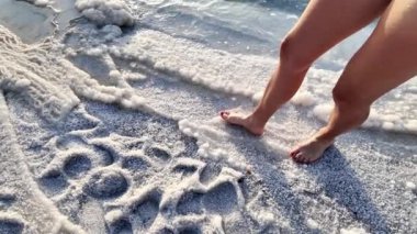 Ürdün 'de tuzla kaplı eşsiz bir plajda yürüyen yalın ayak bir birey sahil boyunca ayak izleri bırakıyor ve dalgalar yavaşça içeri dalıyor.