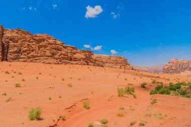 Ürdün 'ün Wadi Rum kentindeki çarpıcı çöl manzarasının panoramik görüntüsü, geniş kumlu bölgeleri ve parlak mavi gökyüzünün altında yükselen kaya oluşumlarını gösteriyor.