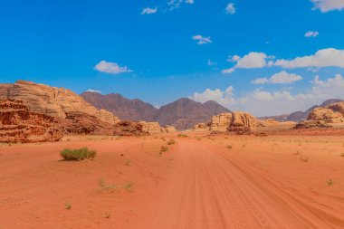 Ürdün 'ün Wadi Rum kentindeki çarpıcı çöl manzarasının panoramik görüntüsü, geniş kumlu bölgeleri ve parlak mavi gökyüzünün altında yükselen kaya oluşumlarını gösteriyor.