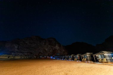 Ürdün 'ün Wadi Rum şehrinde Nisan ayında geleneksel çöl kampı konaklamaları tarafından çerçevelenen eşsiz Jabal al al qattar kaya oluşumunun üzerinde yıldızlarla dolu sakin bir gece.