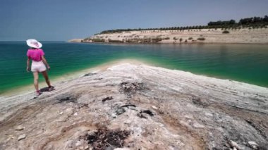 Pembe ve beyaz renkli genç bir kadın, Ürdün 'de berrak, sakin bir gökyüzünün altında Ölü Deniz' in berrak yeşil sularıyla çakıllı bir sahil boyunca yürüyor.