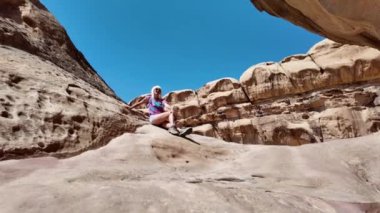 Kadın turist kaşifi Ürdün 'deki Wadi Rum çölünün görkemli kumtaşı oluşumlarıyla çevrili ikonik Um Fruth kaya kemeri köprüsü altında dinleniyor.