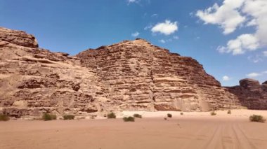 Dalgalı 4x4 araç Wadi Rum Çölü 'nün engin kumlarını aşıyor. Ürdünlüleri sergiliyor doğal güzellikleri ve kayalık araziyi sergiliyor. Parlak mavi gökyüzü altında dağınık bulutlar altında.