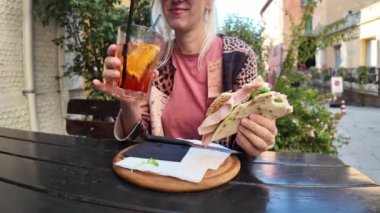 Masada oturan sarışın turist elinde piadina romagnola ve spritz kokteyli tutarak Brisighella 'da tipik bir İtalyan yemeğinin tadını çıkarıyor.