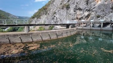 Matka Kanyonu 'nun yapay gölünün etrafındaki beton duvar, su yüzeyinde yüzen yapraklar ve molozlar ve üzerinde metal bir patika var.