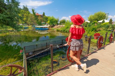 Tourist in a red shirt and wide brimmed hat walks on a wooden bridge with old wagon wheels in a lush park setting of Black Drin springs in lake Ohrid, North Macedonia clipart