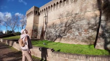 Emilia Romagna 'daki ortaçağ kalesi Dozza' daki Rocca Sforzesca yakınlarında yürüyen sarışın turist güneşli bir kış gününde antik duvarlara ve mavi gökyüzüne hayran.