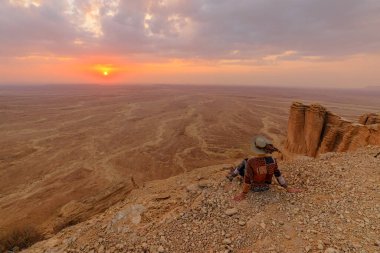Gün batımında dünyanın yamacında oturan şapkalı kadın turist, Suudi Arabistan 'ın Riyad kentindeki Tuwaiq dağ sırasının kayalıklarını inceliyor.