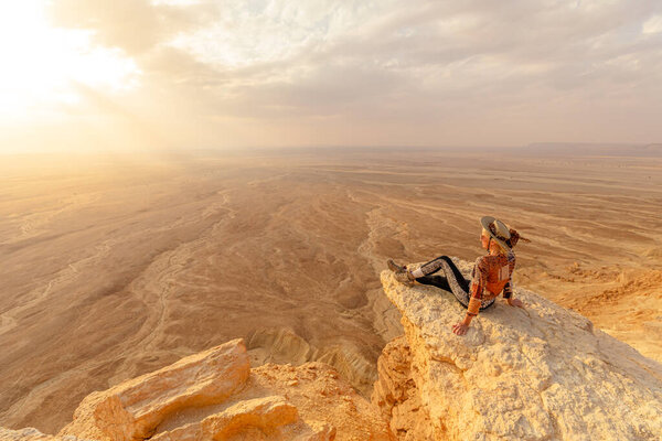 Female tourist wearing hat and sunglasses sitting on rock admiring sunset view of Tuwaiq escarpment, edge of the world, in Riyadh, Saudi Arabia, a breathtaking desert landscape