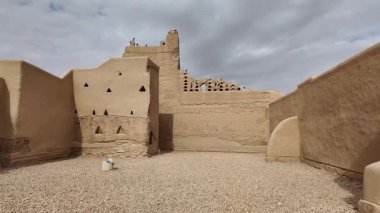 Diriye, Riyad 'daki Suudi Arabistan' ın geleneksel Najdi mimarisi, bulutlu bir gökyüzü altında tarihi koruma ve modern konforların harmanlandığı, dekoratif beyaz süslemeli çamur tuğlalı binalara yer veriyor.