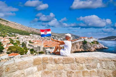 Hırvatistan 'daki Dubrovnik Kalesi' nin tepesinde Hırvat bayrağı taşıyan kadın. Batı limanı manzarası ve şehir duvarları. UNESCO şehri Dubrovnik, Dalmaçya 'nın eski bir Venedik kenti olan Hırvatistan' dır..