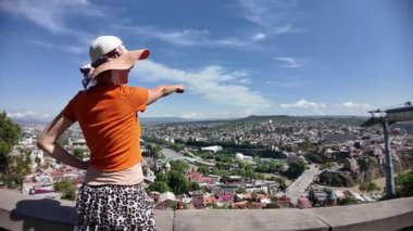 Turuncu tişört ve şapka giyen turist kadın, güzel bir yaz gününde Gürcistan 'da panoramik bir perspektiften görülebilen Kura Nehri ve teleferikli Tiflis şehrine bakıyor.