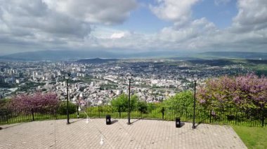 Tiflis 'in verimli yeşillik çerçevelemesi geleneksel ve modern mimarinin bir karışımını ortaya çıkarırken, Kura Nehri güneşli bir gökyüzünün altında canlı şehir manzarası boyunca kıvrılıyor.