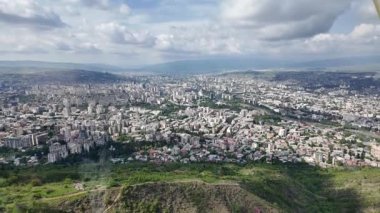Gürcistan 'ın Tiflis kentinin nefes kesici manzarası, yemyeşil ve bulutlu bir gökyüzünün altında yuvarlanan tepelerin ortasında modern ve geleneksel mimarinin bir karışımını gösteriyor.