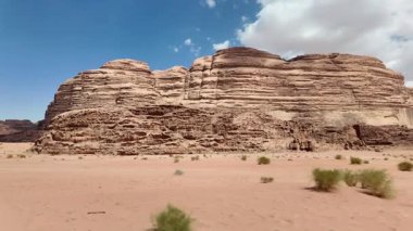 Ürdün 'ün Wadi Rum kentindeki çarpıcı çöl manzarasının panoramik görüntüsü, geniş kumlu bölgeleri ve parlak mavi gökyüzünün altında yükselen kaya oluşumlarını gösteriyor.