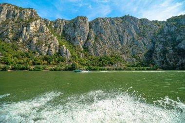 Demir Kapılar geçidinde Tuna Nehri 'nden geçen sürat teknesi, sudan dik bir şekilde yükselen orman dağları ve Romanya kıyısı boyunca yayılmış bir köyle