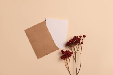 Mockup of an open invitation card made of eco-friendly kraft paper and decorated with a composition of dried flowers painted in burgundy color, arranged vertically on a beige background