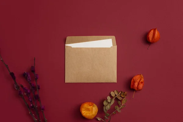 Flatlay mockup of a card with three orange physalis flowers and a composition of dry branches on kraft paper against a burgundy background