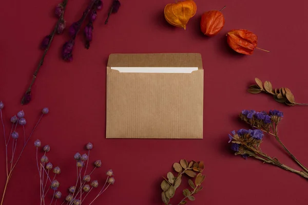 Flatlay mockup of a card made of kraft paper with a composition of dried flowers, physalis, and painted branches on a burgundy background