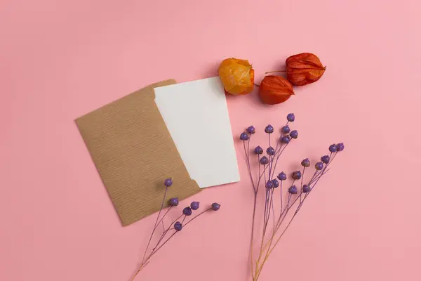 Flatlay mockup of a greeting card made of craft paper with three orange physalis flowers and a composition of dry twigs on a pink background