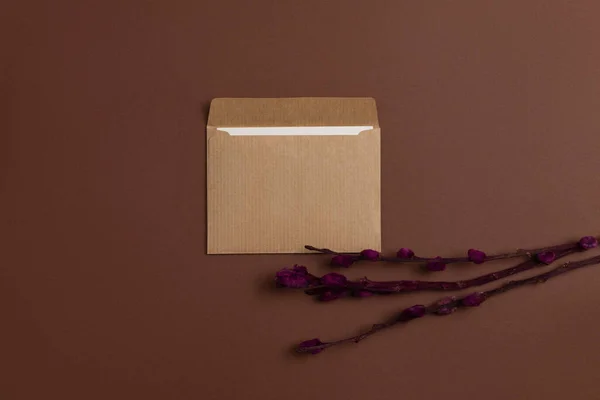 Mockup of a card made from eco-friendly kraft paper and decorated with a composition of dried blooming branches painted in burgundy color and placed on a horizontal brown background