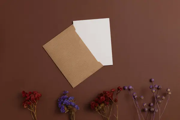 Open invitation card made of eco-friendly kraft paper and decorated with a composition of dry flowers in red and purple colors, placed at the bottom on a brown background