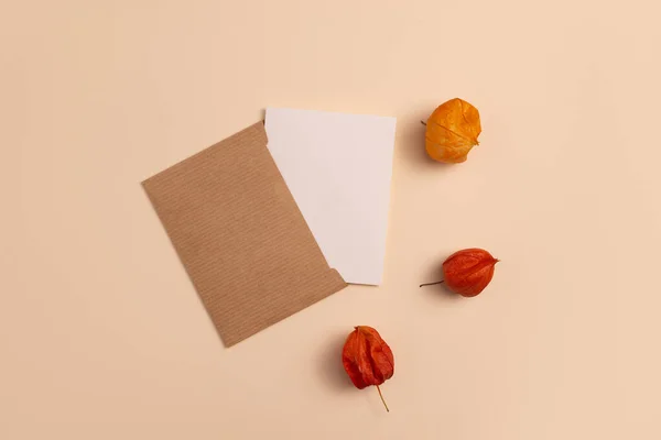 Mockup of an open invitation card made of ecological kraft paper and decorated with a composition of orange and red Physalis flowers placed on a beige background