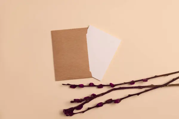 Mockup of an open invitation card made from eco-friendly kraft paper and decorated with a composition of dried flowering branches painted in burgundy color, arranged horizontally on a beige background