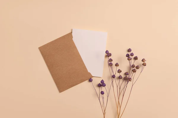 Mockup of an open invitation card made of eco-friendly craft paper and decorated with a composition of dried flax flowers painted in purple, arranged vertically on a beige background