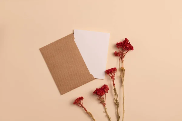 Mockup of an open invitation card made of ecological kraft paper and decorated with a composition of dried flowers painted in red color, arranged vertically on a beige background