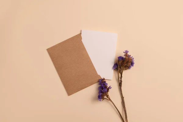 Mockup of an open invitation card made of eco-friendly kraft paper and decorated with a composition of blue dried flowers, arranged vertically on a beige background