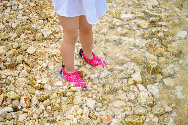 Childrens feet in bright aqua shoes in the water on a rocky bottom.