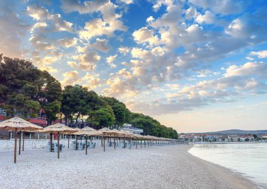 Şafakta saman şemsiyeli plaj. Çakıl taşlarını ve şehrin bir kısmını görebilirsin..