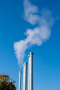 Several large metal pipes produce white steam against the background of the blue sky. Pipes of the heating boiler that gives heat to the houses. clipart