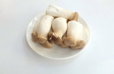 Üst ve düz bir beyaz arka planda izole edilmiş beyaz bir tabakta çiğ çiğ kral istiridye mantarları ve rosmary (Pleurotus eryngii) yatar.