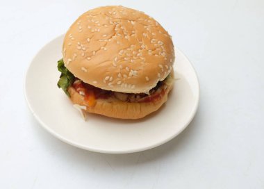 Domuz pirzolası ve domatesli klasik burger. Salatalık, kırmızı soğan, peynir ve marullu susamlı çörek. Beyaz bir arka planda izole edilmiş beyaz plaka üzerinde. Amerikan hamburgeri
