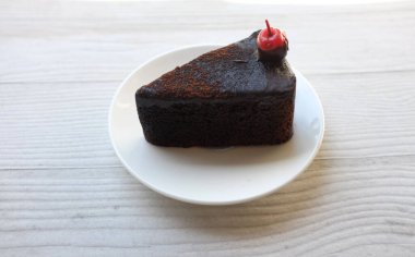 Beyaz tabakta kırmızı kirazla süslenmiş katmanlı siyah çikolatalı pasta beyaz ahşap arka planda izole edilmiş.