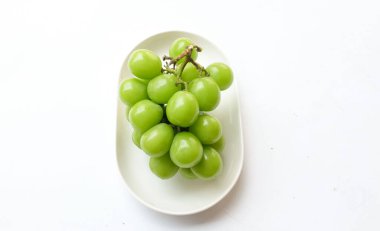 Beyaz bir sırt damlasında beyaz oval seramik tabakta izole edilmiş taze, tatlı yeşil Muscat (Vitis vinifera) üzümü. Japon üzümleri. Üzüm yeşili. Tadı tatlı ve doğal..