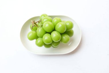 Beyaz bir sırt damlasında beyaz oval seramik tabakta izole edilmiş taze, tatlı yeşil Muscat (Vitis vinifera) üzümü. Japon üzümleri. Üzüm yeşili. Tadı tatlı ve doğal..