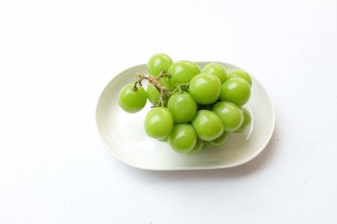 Beyaz bir sırt damlasında beyaz oval seramik tabakta izole edilmiş taze, tatlı yeşil Muscat (Vitis vinifera) üzümü. Japon üzümleri. Üzüm yeşili. Tadı tatlı ve doğal..