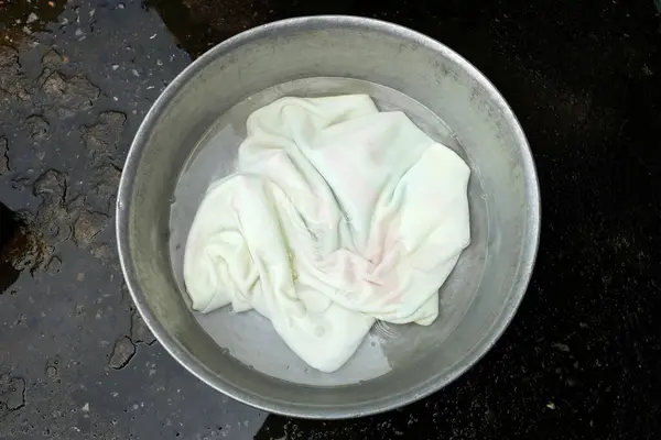 A white towel is soaked in an aluminum basin with detergent to prepare for hand washing.