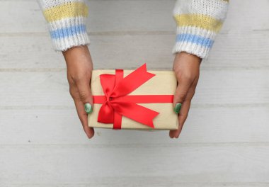 Renkli süveter giymiş Asyalı bir kızın elinde beyaz tahta bir masa üzerinde kırmızı kurdele bulunan el işi bir hediye paketi var. Özel bir gün, Noel, yeni yıl, sevgililer günü için.