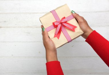 Renkli süveter giymiş Asyalı bir kızın elinde beyaz tahta bir masa üzerinde kırmızı kurdele bulunan el işi bir hediye paketi var. Özel bir gün, Noel, yeni yıl, sevgililer günü için.